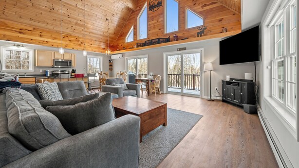 A spacious living room with a high ceiling and wooden beams, a gray sofa, a wooden coffee table, and a TV mounted on the wall. - Deer Crossing Retreat - new home close to Saddleback (Dallas Plantation)