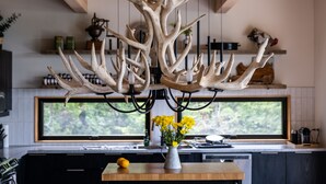 Una cocina moderna con una lámpara original hecha de astas de ciervo, una mesa de madera con un jarrón de flores amarillas, y un amplio ventanal con vistas a zonas verdes.