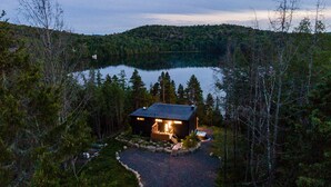 Una cabaña con porche y una pequeña terraza, rodeada de un bosque denso y un lago.
