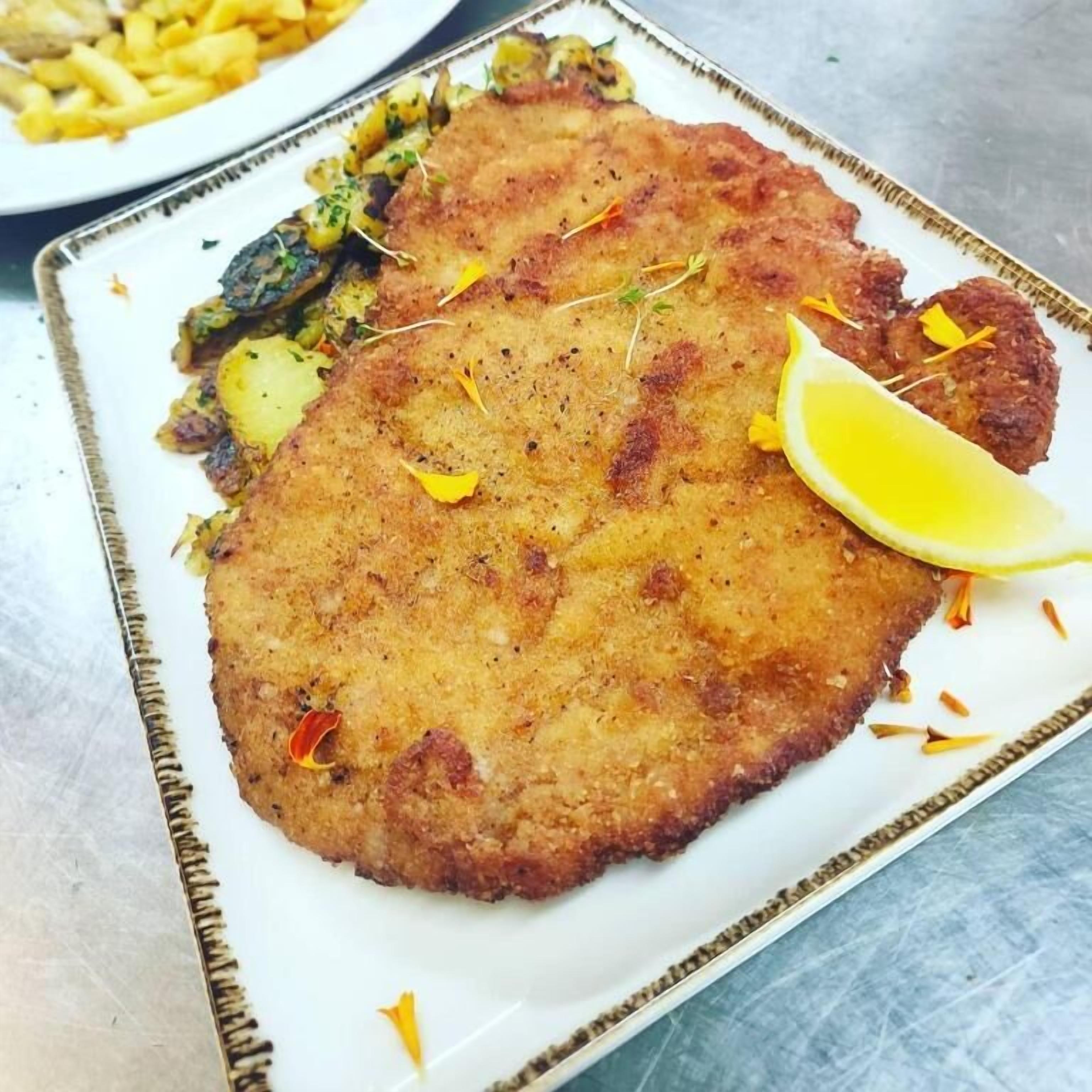 Un plato con una milanesa cubierta de pan rallado, adornada con limón y hierbas, acompañada de papas fritas y verduras.
