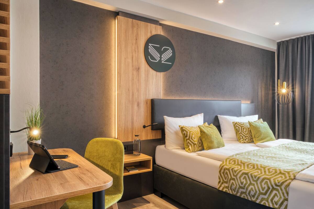 Une chambre d’hôtel moderne, équipée d’un lit, d’un bureau avec un ordinateur portable, d’un fauteuil vert et d’un panneau indiquant le numéro de la chambre, fixé au mur.