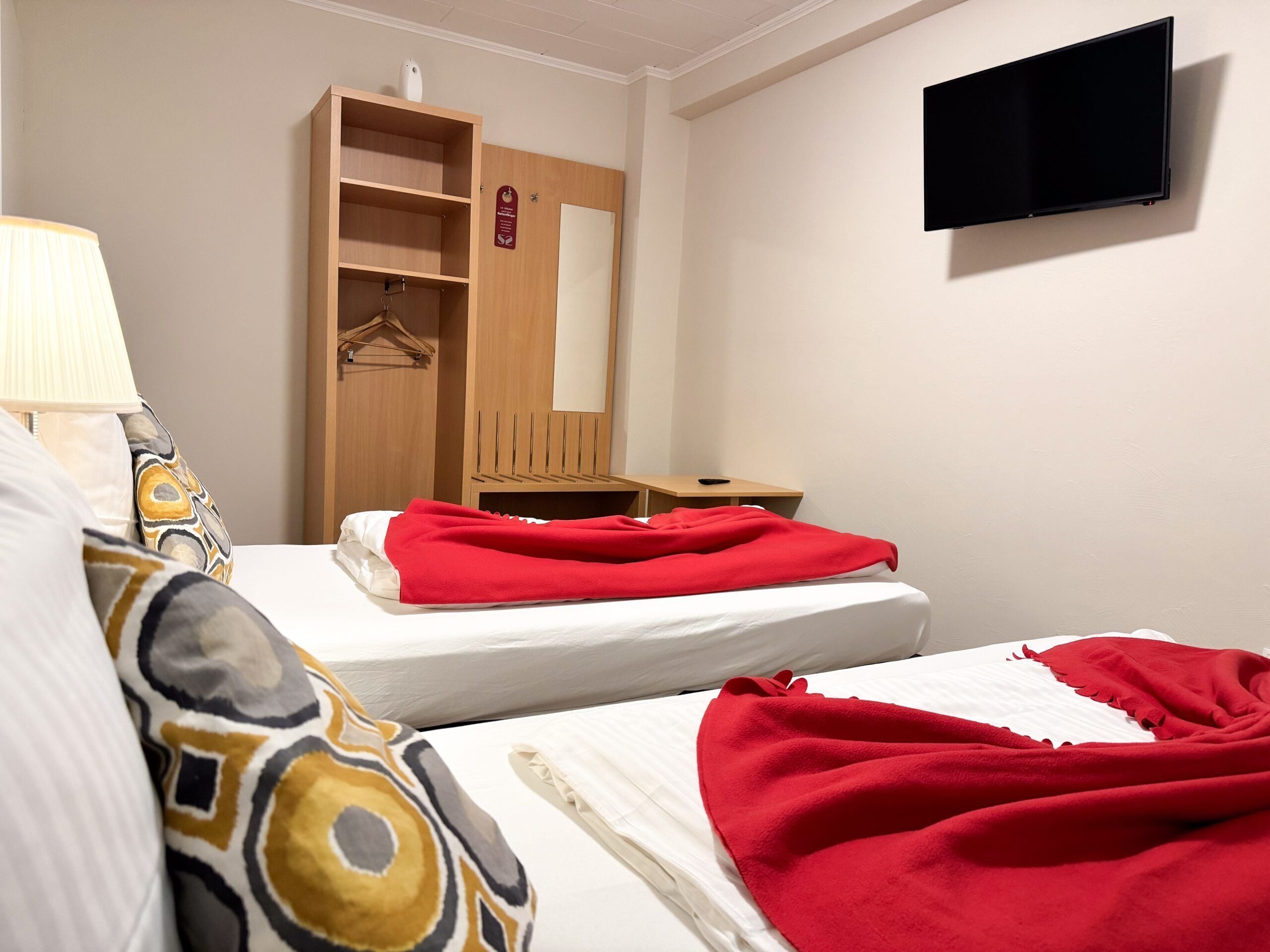 Habitación de hotel con dos camas, un armario de madera, una mesita de noche, un televisor montado en la pared y una lámpara.