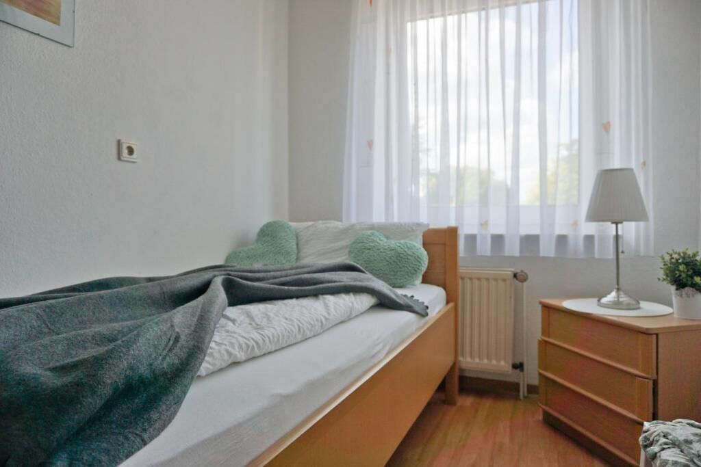 A bedroom with a bed, a bedside table with a lamp, a radiator, and a window with sheer curtains.