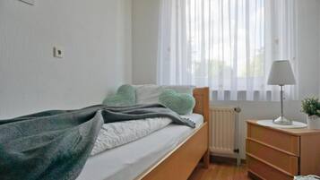 A bedroom with a bed, a bedside table with a lamp, a radiator, and a window with sheer curtains.