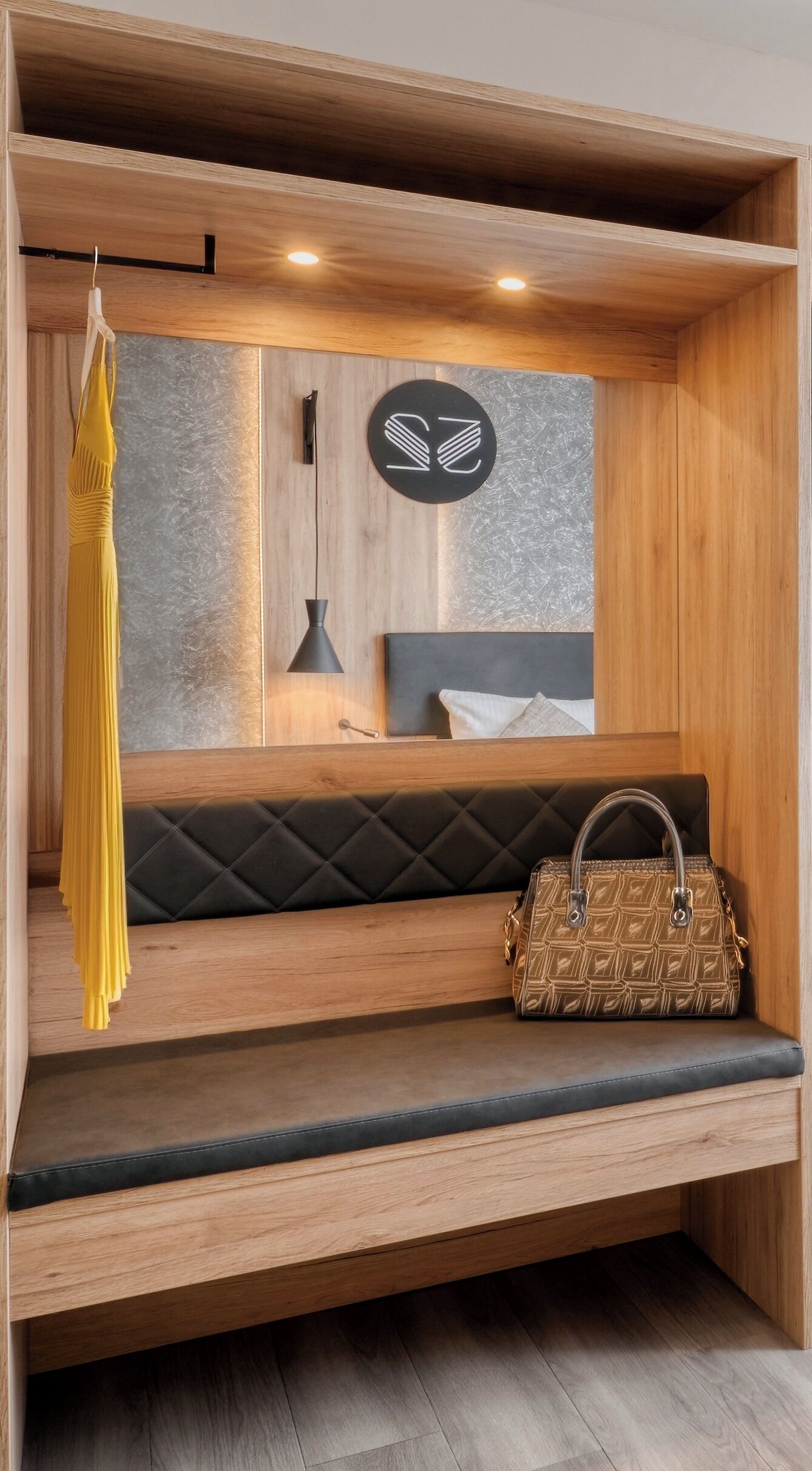 A wooden bench with a cushioned seat, a hanging yellow garment, a wall-mounted black logo, and a patterned handbag on the bench.