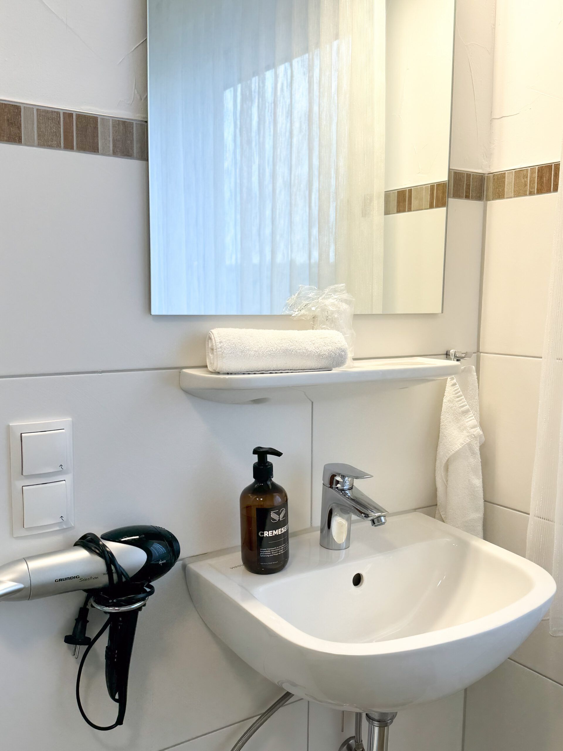Un bagno con un lavandino bianco, uno specchio, un asciugamano, una crema e un asciugacapelli.