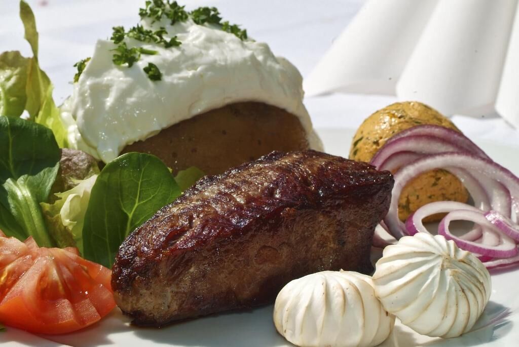 Un piatto con una bistecca alla griglia, un patate al forno con panna acida ed erbe aromatiche, un pomodoro alla griglia, cipolle rosse a fette e spicchi d'aglio.