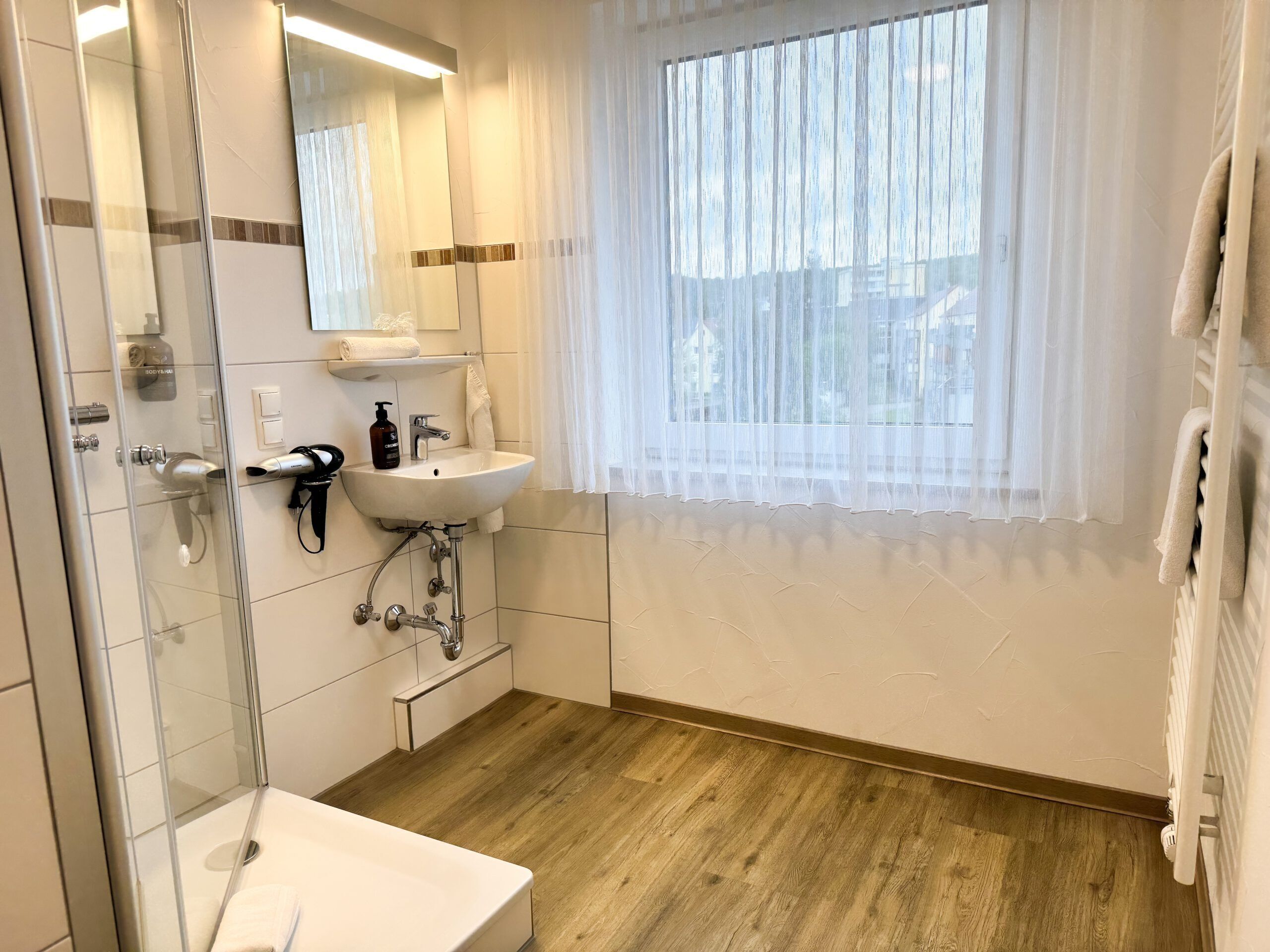 A modern bathroom with a glass shower enclosure, a white sink, and a window with sheer curtains.