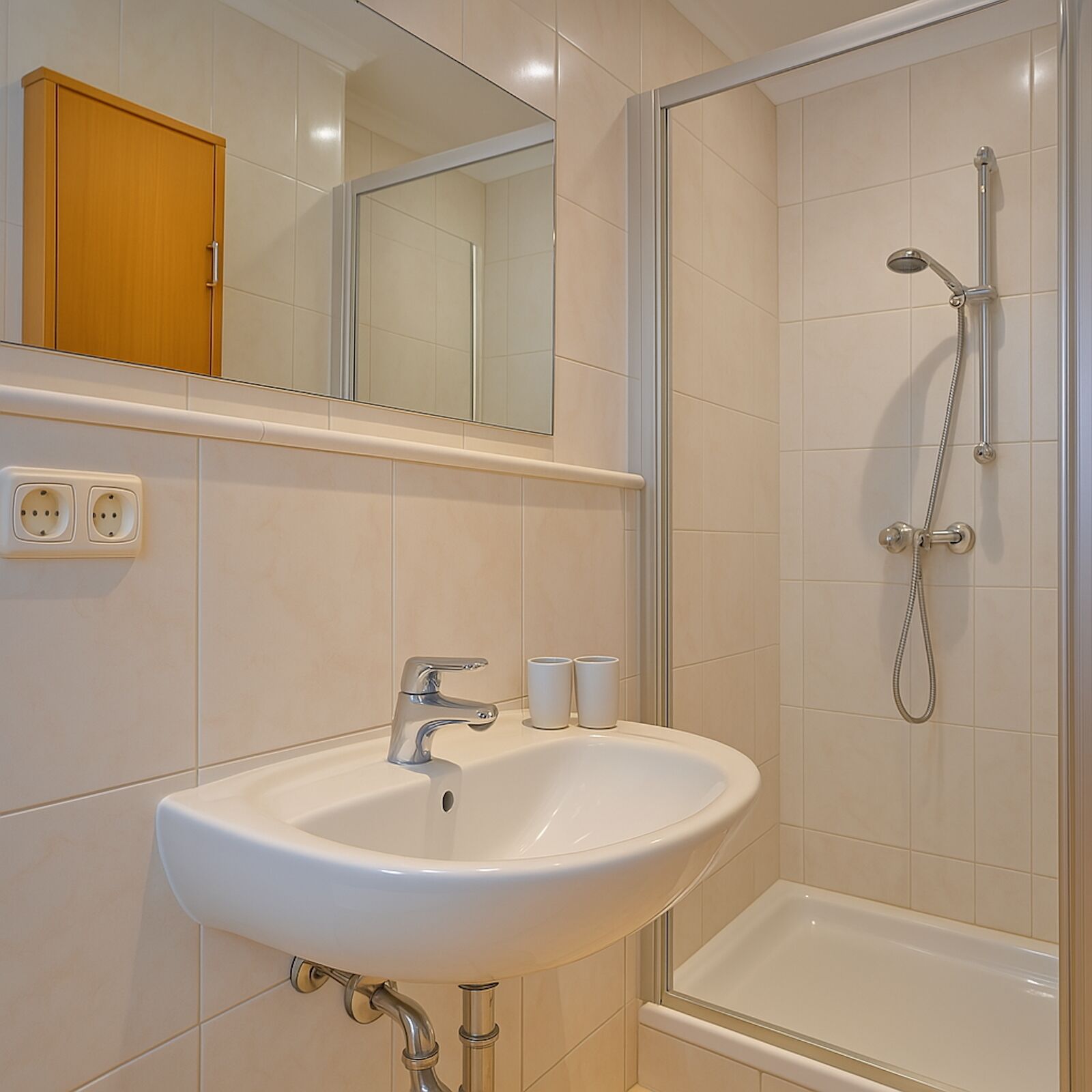 Un baño con lavamanos blanco, un espejo, ducha y un armario de madera.