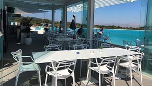 Un área de comedor al aire libre con sillas y mesas blancas, con vistas a un mar azul cristalino y una costa distante.