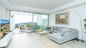 Una sala moderna con un sofá gris, una mesa de centro y un televisor sobre un soporte blanco. Se ve un amplio ventanal con vistas a un paisaje verde.