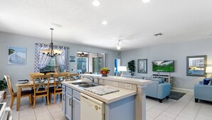 A modern kitchen with a dishwasher, a dining area with wooden chairs, a blue sofa, and a TV mounted on the wall.