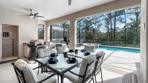 A spacious outdoor dining area with a glass table and chairs, a ceiling fan, and a view of a swimming pool and trees.