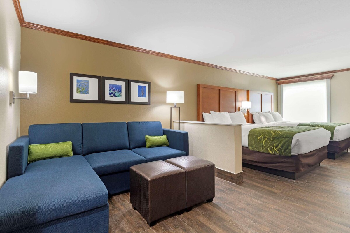 A hotel room with a blue sofa, a small ottoman, a bed with a green blanket, and framed pictures on the wall.