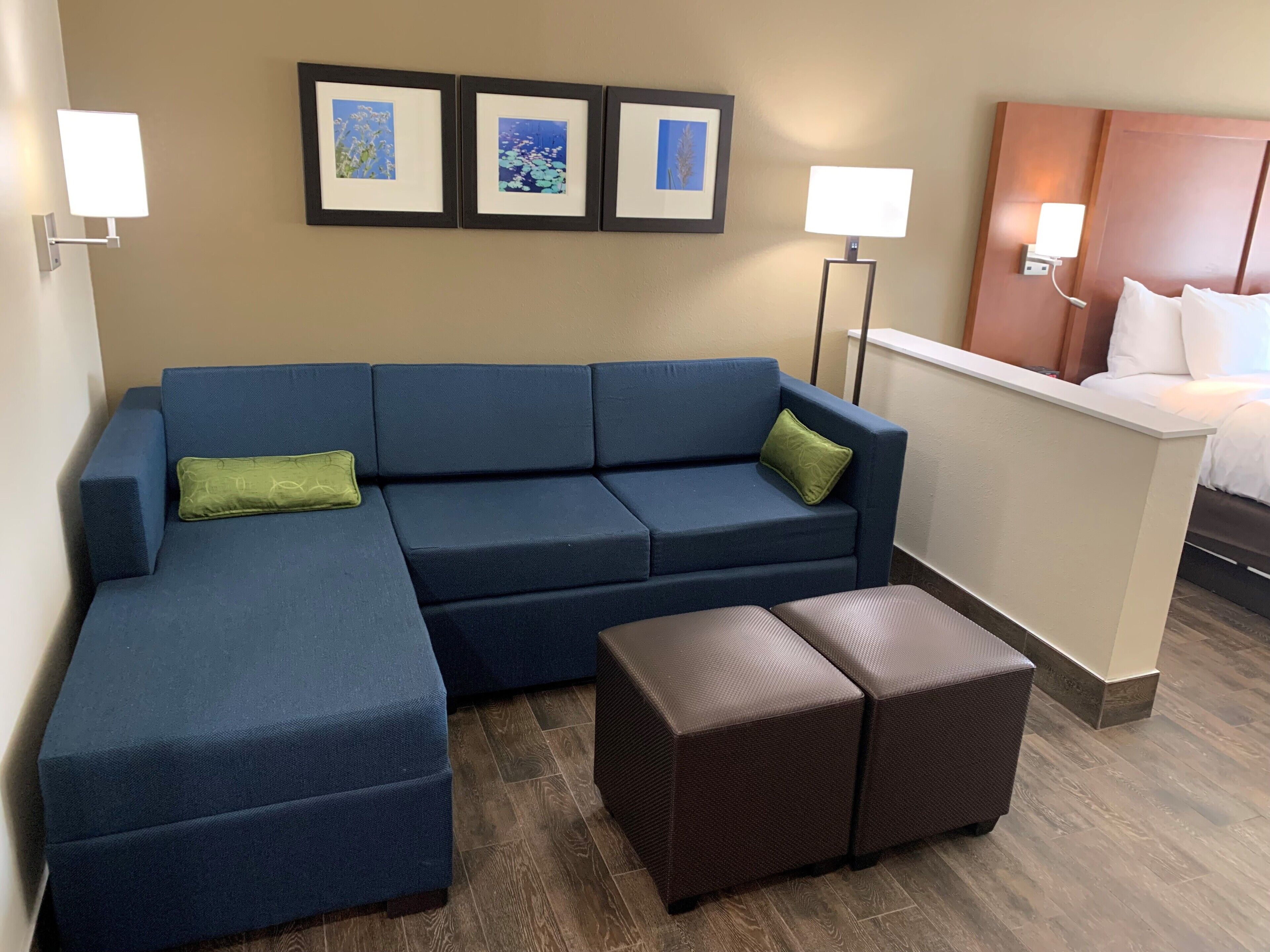 A hotel room with a blue sectional sofa, two green pillows, a brown ottoman, and a bed with white linens. There are three framed pictures on the wall and two wall-mounted lamps.