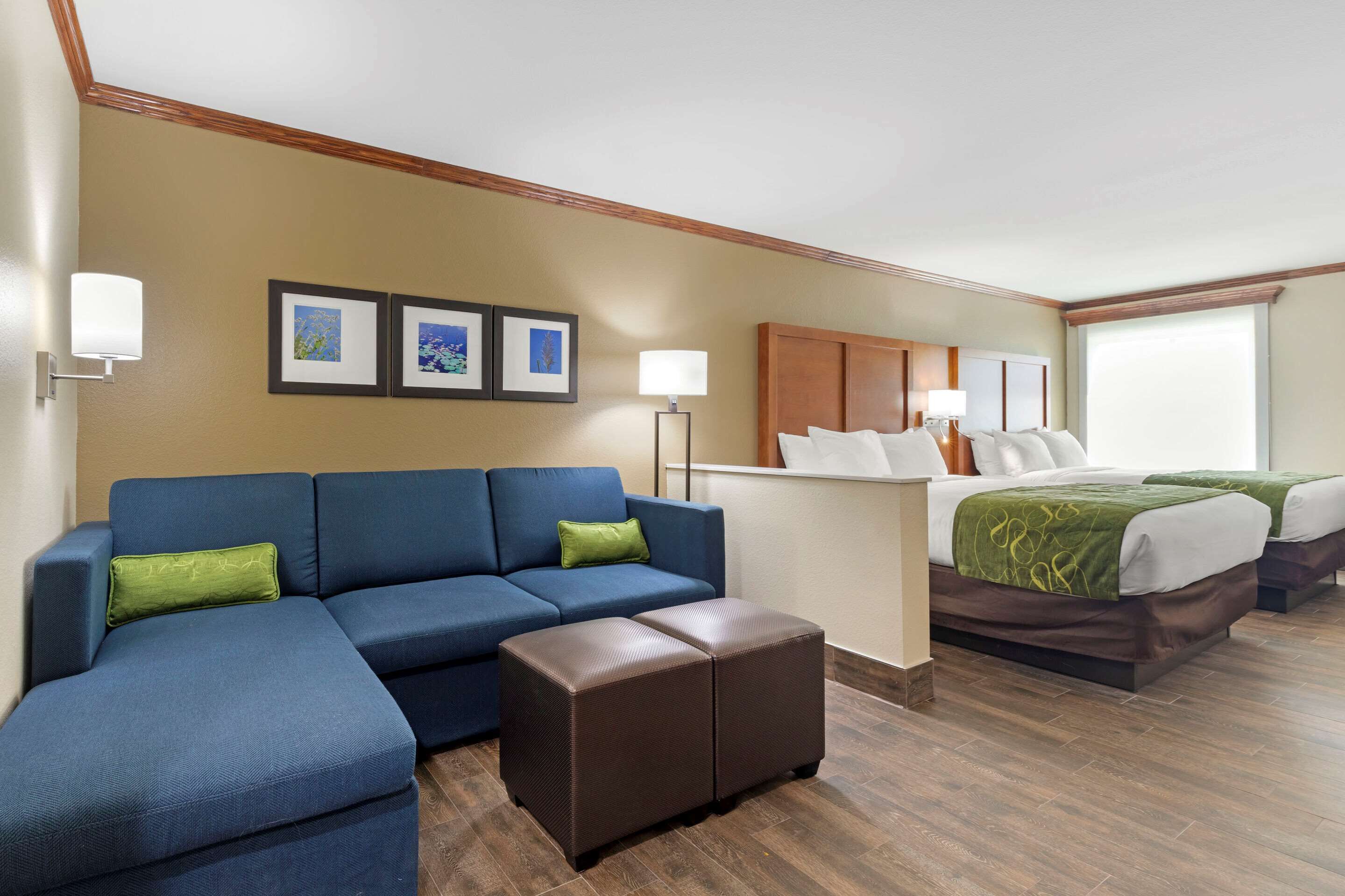 Habitación de hotel con un sofá azul, un pequeño reposapiés, una cama con ropa de cama verde y blanca y un cabecero de madera. Hay tres cuadros enmarcados en la pared y una lámpara de pie.