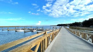 A wooden pier extending into calm water with boats docked, a clear sky, and a distant shoreline with trees and buildings. - Coastal stay/steps to Driftwood Beach (Jekyll Island)