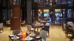 Un restaurant moderne avec des tables dressées pour le service, un serveur qui s’affaire et une vue sur la cuisine à travers de grandes fenêtres.