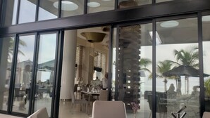 Un hall d’hôtel moderne avec de grandes portes vitrées, un espace repas avec des tables et des chaises, et une vue sur les palmiers et la mer.