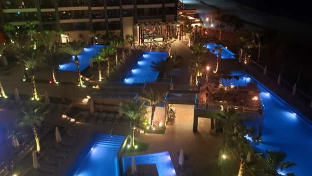 Vue nocturne d’un espace piscine d’hôtel, avec plusieurs piscines, des palmiers et un éclairage d’ambiance.
