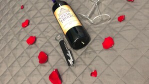 Una botella de vino, un sacacorchos y dos copas de vino sobre una superficie acolchada con pétalos de rosa dispersos.