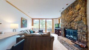 A living room with a stone fireplace, a sofa, a TV, and a large window. - Villas at Snowmass Club - 1422 ~ Snowmass Club Access ~ (Snowmass Village)