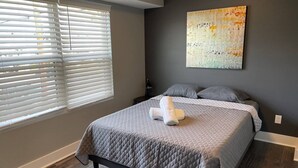 Un dormitorio con cama, una ventana con persianas, una mesita de noche, una lámpara y un cuadro en la pared.