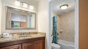 Een badkamer met een granieten aanrechtblad, houten kastjes, een grote spiegel, een toilet en een douche met een blauw gordijn.
