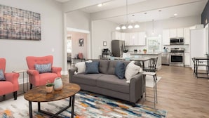 A spacious living room with a grey sofa, red armchairs, and a coffee table. A kitchen with white cabinets and modern appliances is visible in the background. - Charming Modern Farm House-5 min from Sport Center (Rock Hill)