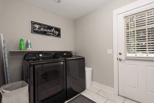 A laundry room with two front-loading washers and dryers, a shelf with laundry supplies, and a door with blinds.