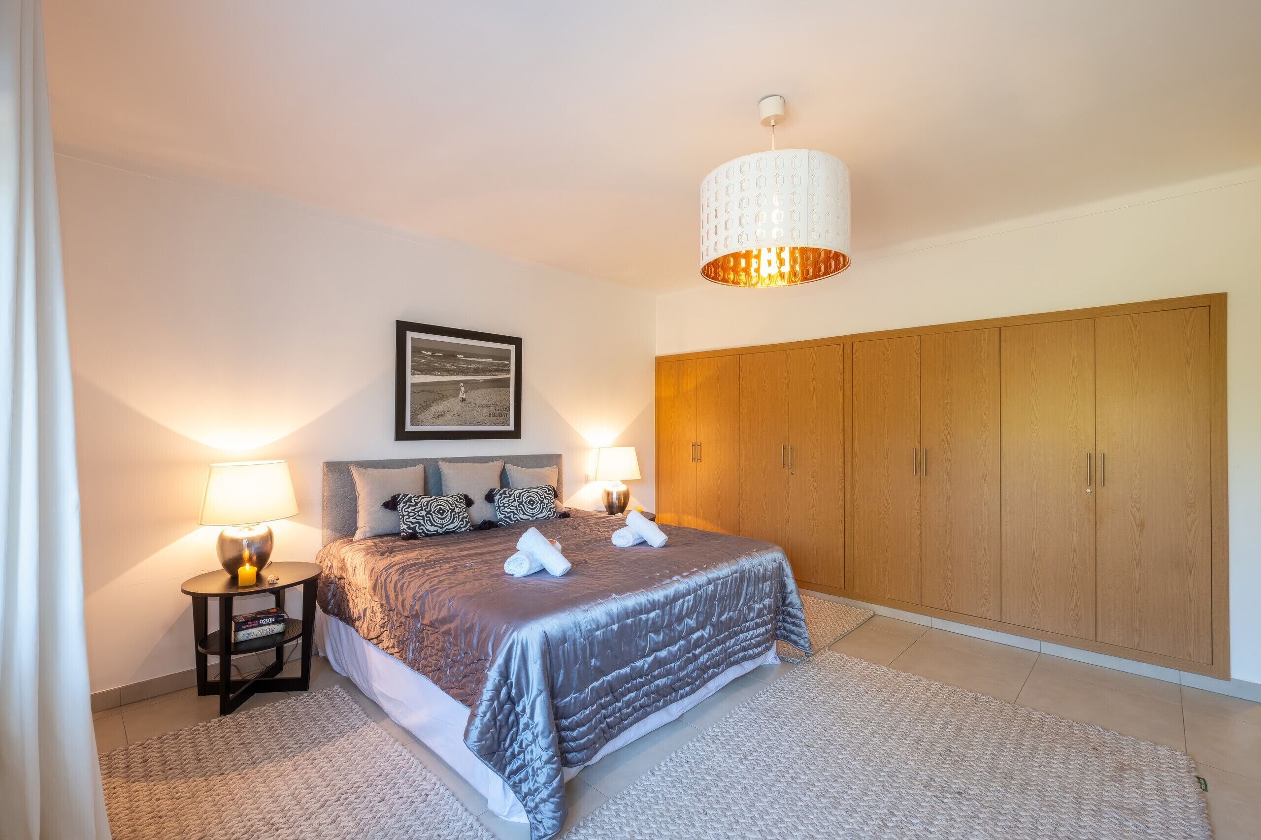 A hotel room with a large bed, two bedside lamps, a framed picture on the wall, a wardrobe, and a hanging light fixture.