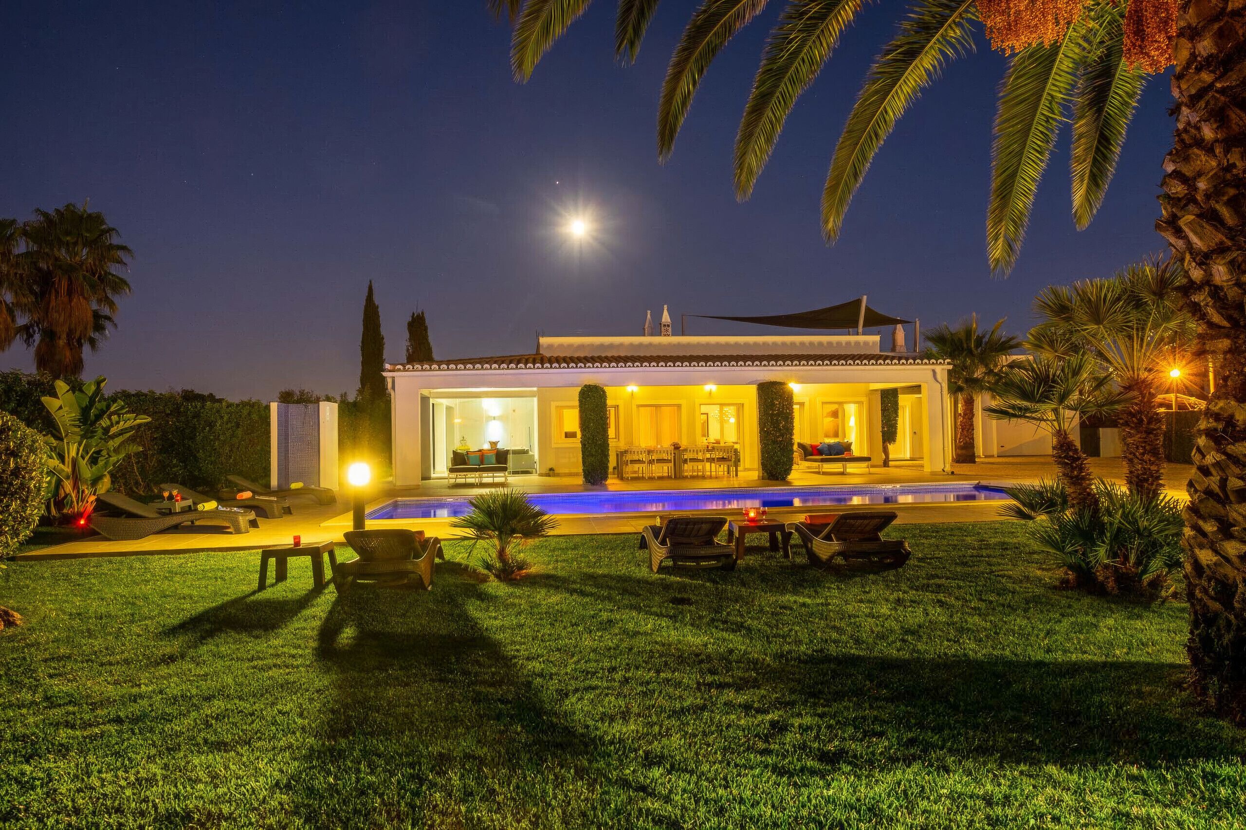 Une belle maison d’habitation, bien éclairée la nuit, avec une piscine, un espace salon extérieur et une végétation luxuriante.