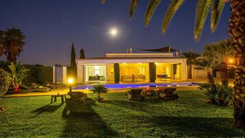 Une belle maison d’habitation, bien éclairée la nuit, avec une piscine, un espace salon extérieur et une végétation luxuriante.