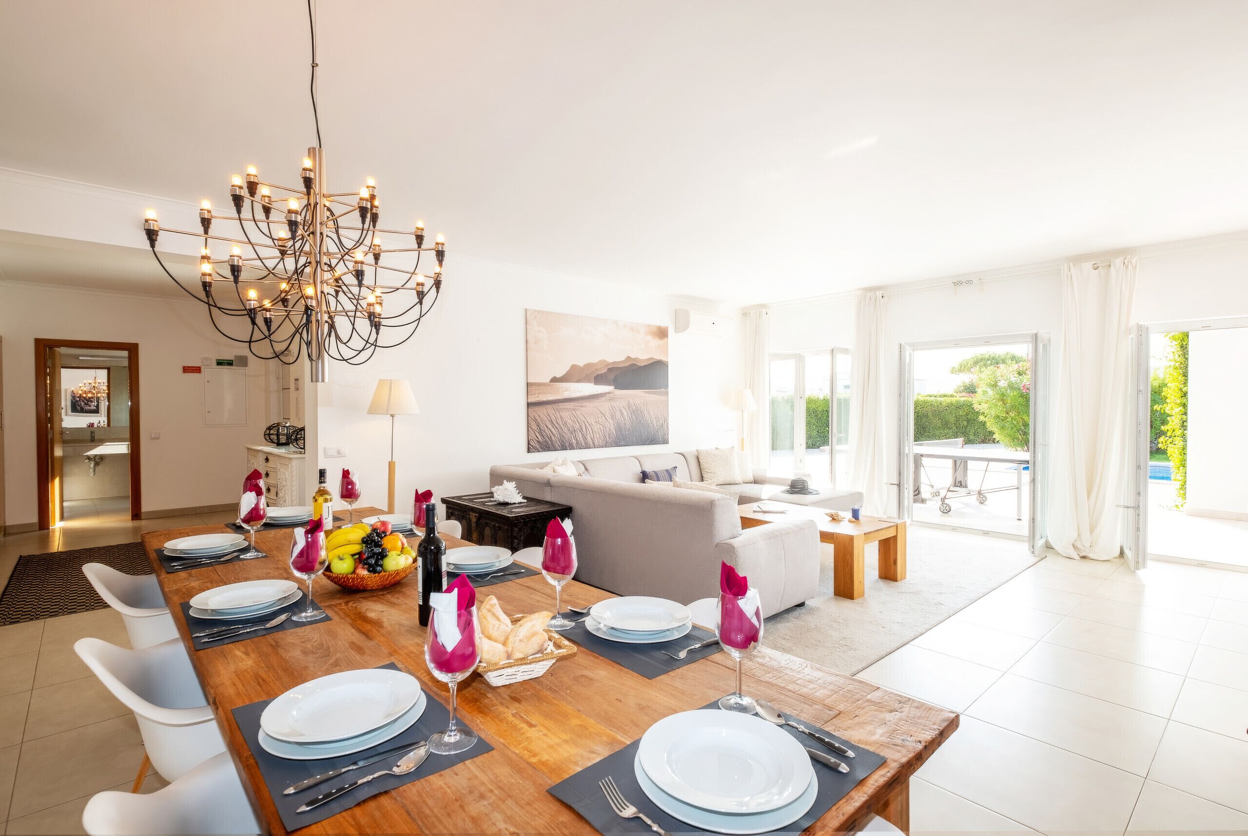 A dining area with a wooden table set for a meal, a chandelier, and a living room with a sofa and a painting.