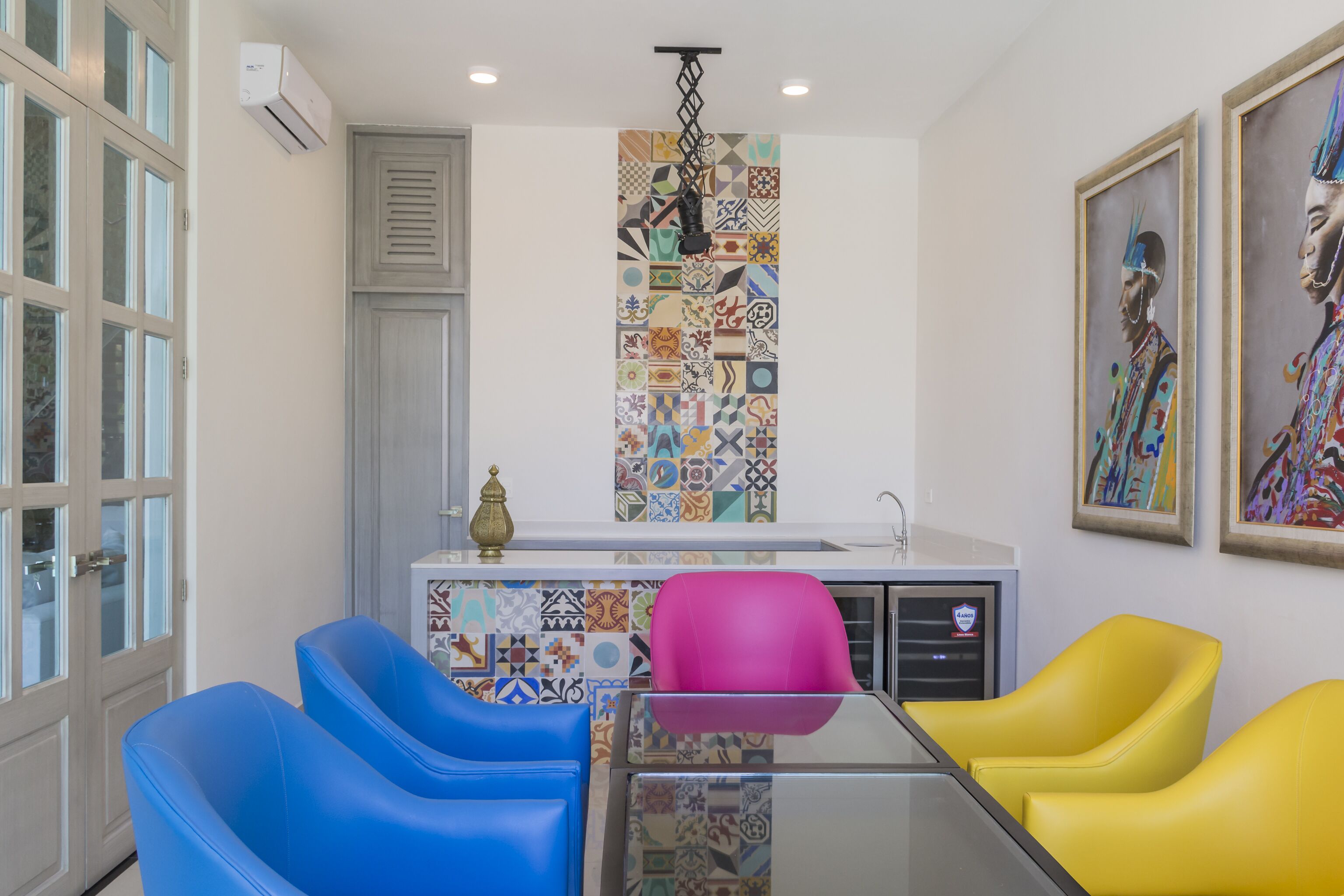 A dining area with a glass table, blue and yellow chairs, a colorful tile wall, and a painting on the wall.