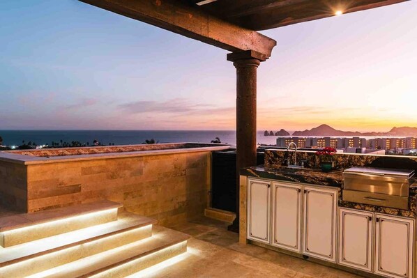 A rooftop bar with steps leading down to the terrace, overlooking a cityscape and the sea at sunset.