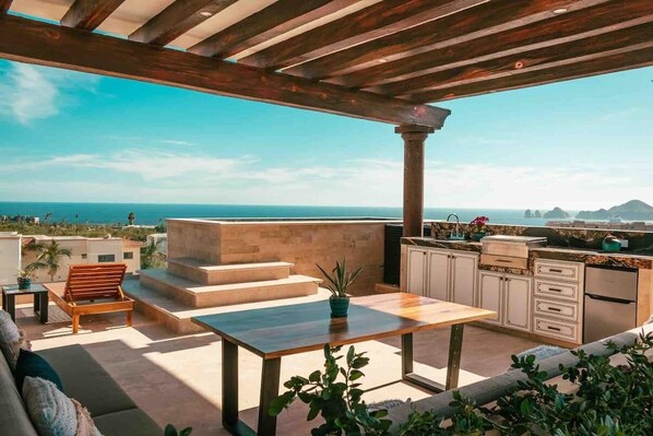 A rooftop terrace with a wooden pergola, a dining table, a sofa, and a kitchen area with a view of the sea.