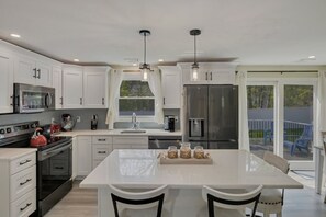 A modern kitchen with white cabinets, stainless steel appliances, a central island with chairs, and a view of the backyard through large windows. - 🐶 Dog Friendly Fenced in Yard! 3/2 🏠 1.1 mi to 🏝 WIFI - perfect fall rental! (Chatham)