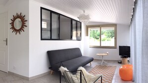 A modern living room with a sofa, a television, a small table, and a window with a view of greenery. - Maisonnette with swimming pool access, on the campsite (Aix en Provence)