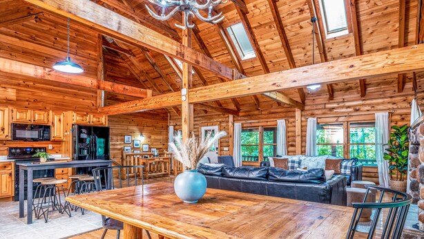 Interior espacioso de una cabaña de madera con una gran mesa de comedor, zona de cocina y un rincón de estar acogedor con un sofá y sillones.