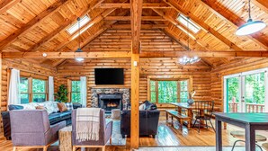Interior de una cabaña de troncos con chimenea, muebles de madera y ventanas amplias.