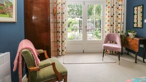 A room with a green chair, a pink chair, a wooden wardrobe, a white radiator, a white ceiling fan, a white door with polka dot curtains, and a view of a garden through the door. - The Barn at Sandhole Cottage (Tonbridge)