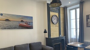 En stue med en grå sofa, et billede af en båd på vandet, en pejs og et spiseområde med et bord og stole.