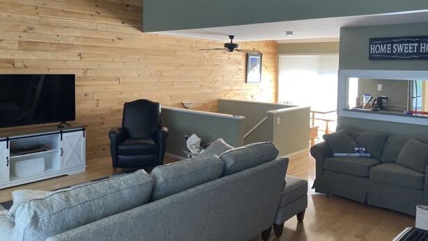Ein Wohnzimmer mit Fernseher, einem schwarzen Sessel, einem grauen Sofa und einem grauen Zweisitzer. An der Decke befindet sich Holz und ein Schild mit der Aufschrift 'HOME SWEET HOME'.
