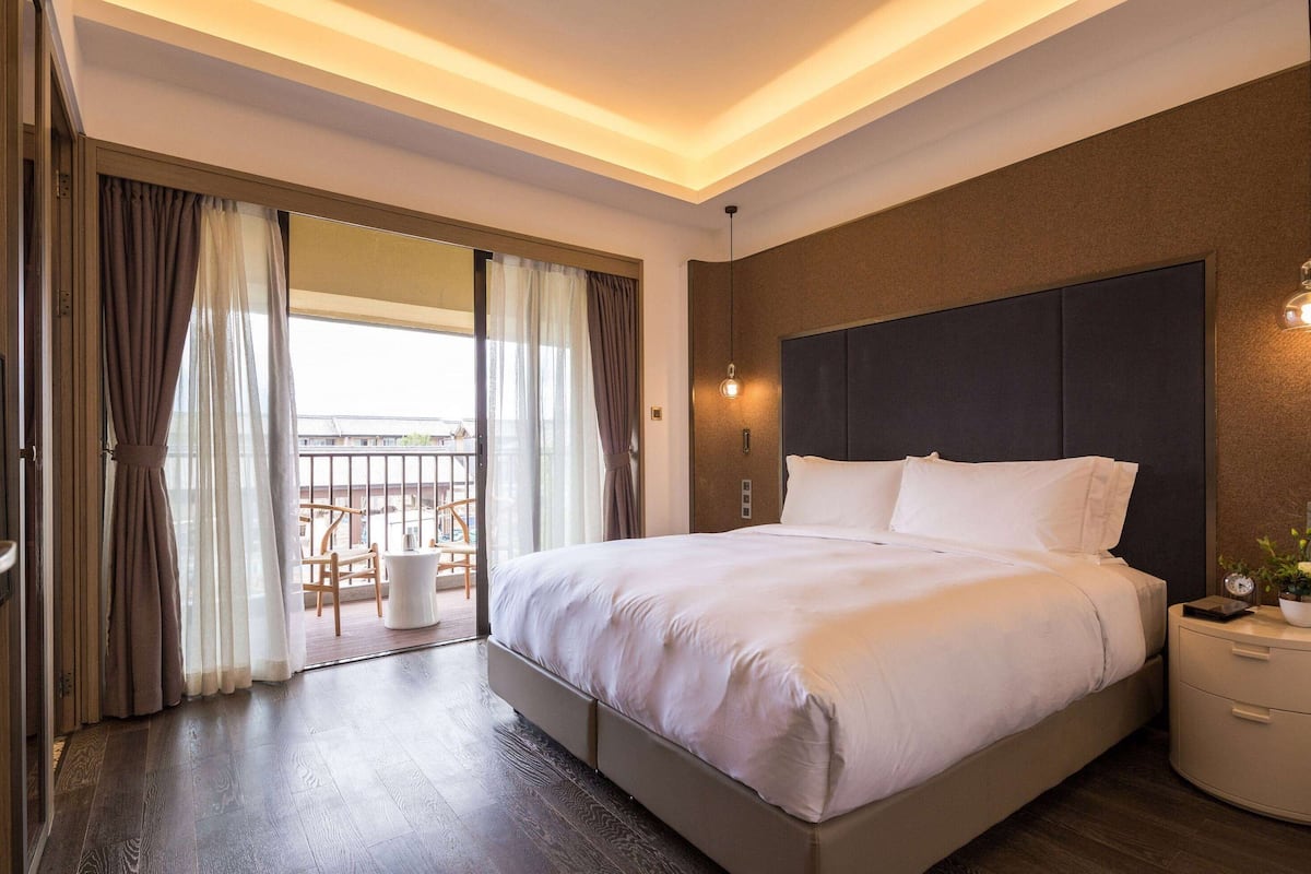 A modern hotel room with a large bed, a bedside table, a nightstand, and a view of a balcony with outdoor furniture.