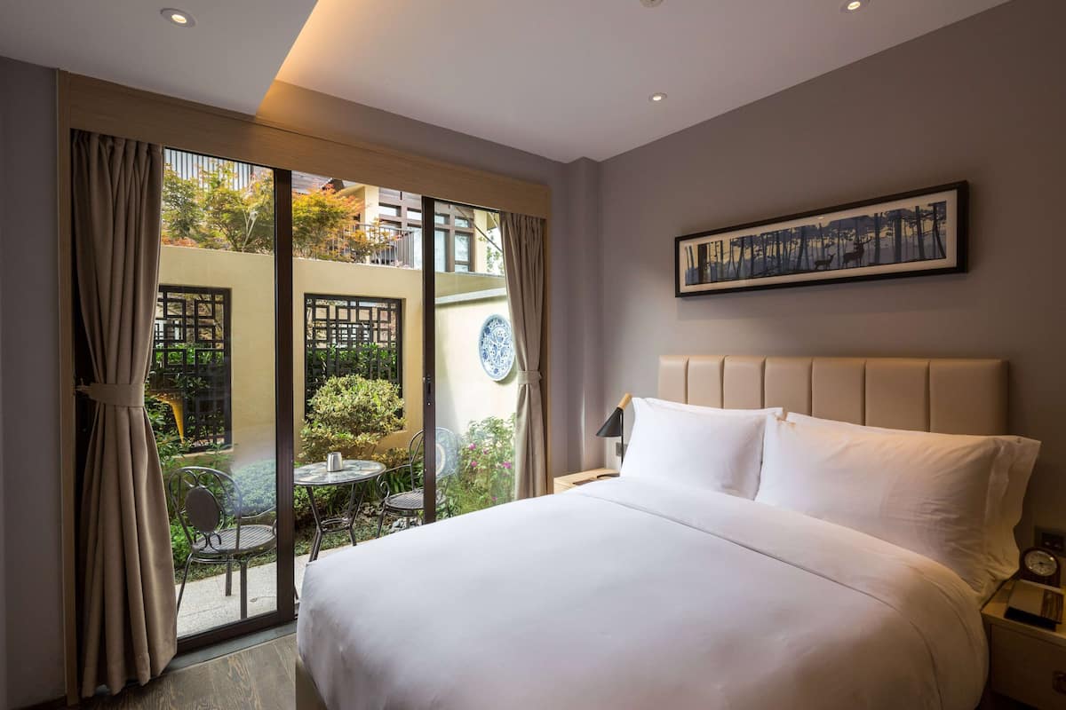A hotel room with a large bed, a view of a garden through a sliding door, a framed artwork on the wall, and a nightstand with a lamp.