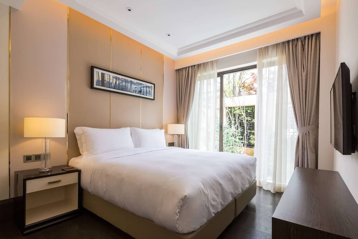 A hotel room with a large bed, two bedside tables with lamps, a flat-screen TV, and a window with curtains.