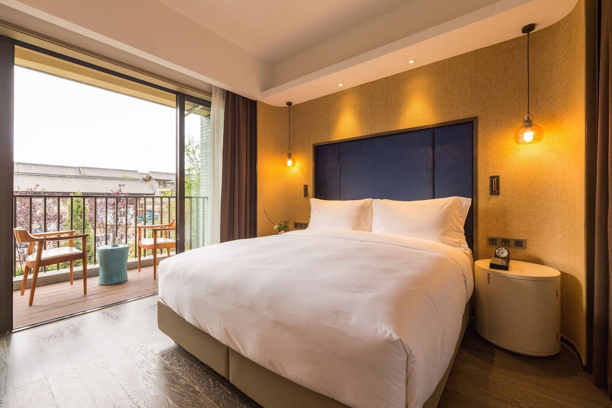 A large bed with a headboard, a bedside table with a clock, and a small round nightstand. There is a window with a view of a balcony and a building outside.