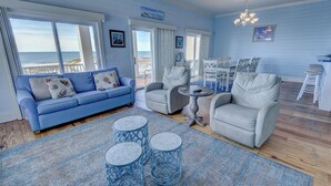 Ein geräumiges Wohnzimmer mit einem blauen Sofa, zwei Sesseln, einem Couchtisch und Blick auf den Strand durch große Fenster.