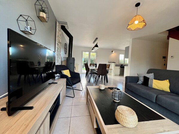 A modern living room with a flat-screen TV, a sofa, a dining area, and a kitchen in the background.